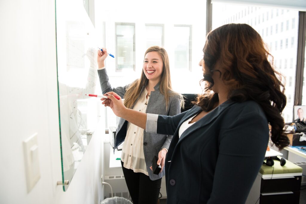 Two women are writing on a whiteboard.